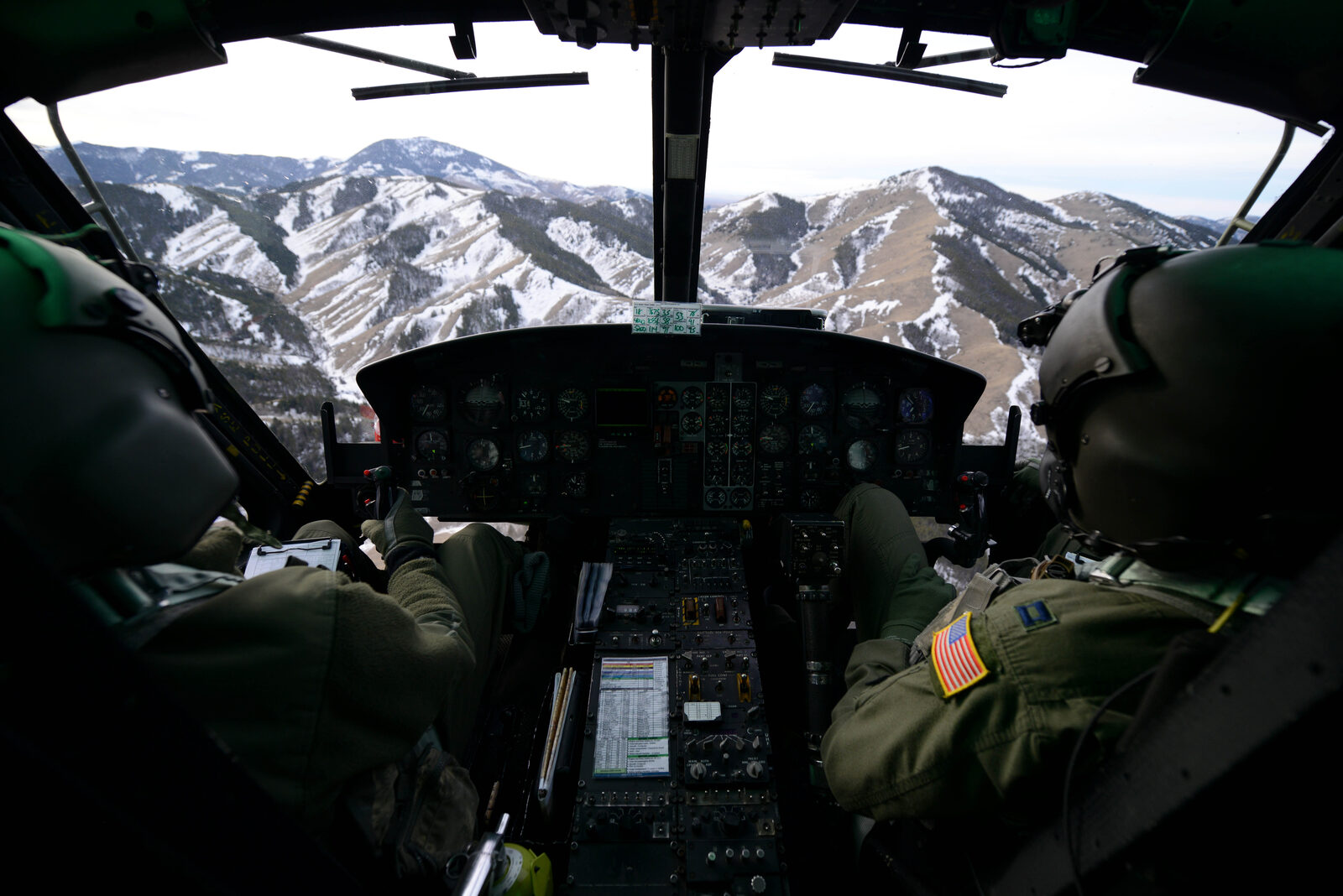 Two Air Force helicopter pilots at the controls of a UH-1N Iroquois, hands on the cyclic and collective, flying over a snow-covered mountain range